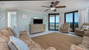 Ein großzügiges Wohnzimmer mit einem großen Flachbildfernseher, einem Deckenventilator und Meerblick durch große Schiebeglastüren.