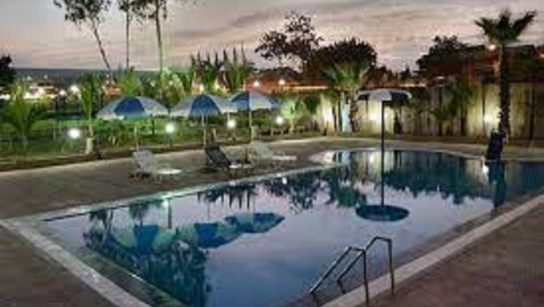 A swimming pool with lounge chairs and umbrellas, surrounded by palm trees and a view of the ocean at dusk. - Hotel Vegas (Khandala)