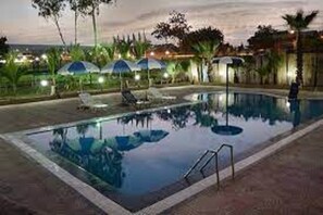 A swimming pool with lounge chairs and umbrellas, surrounded by palm trees and a view of the ocean at dusk. - Hotel Vegas (Khandala)