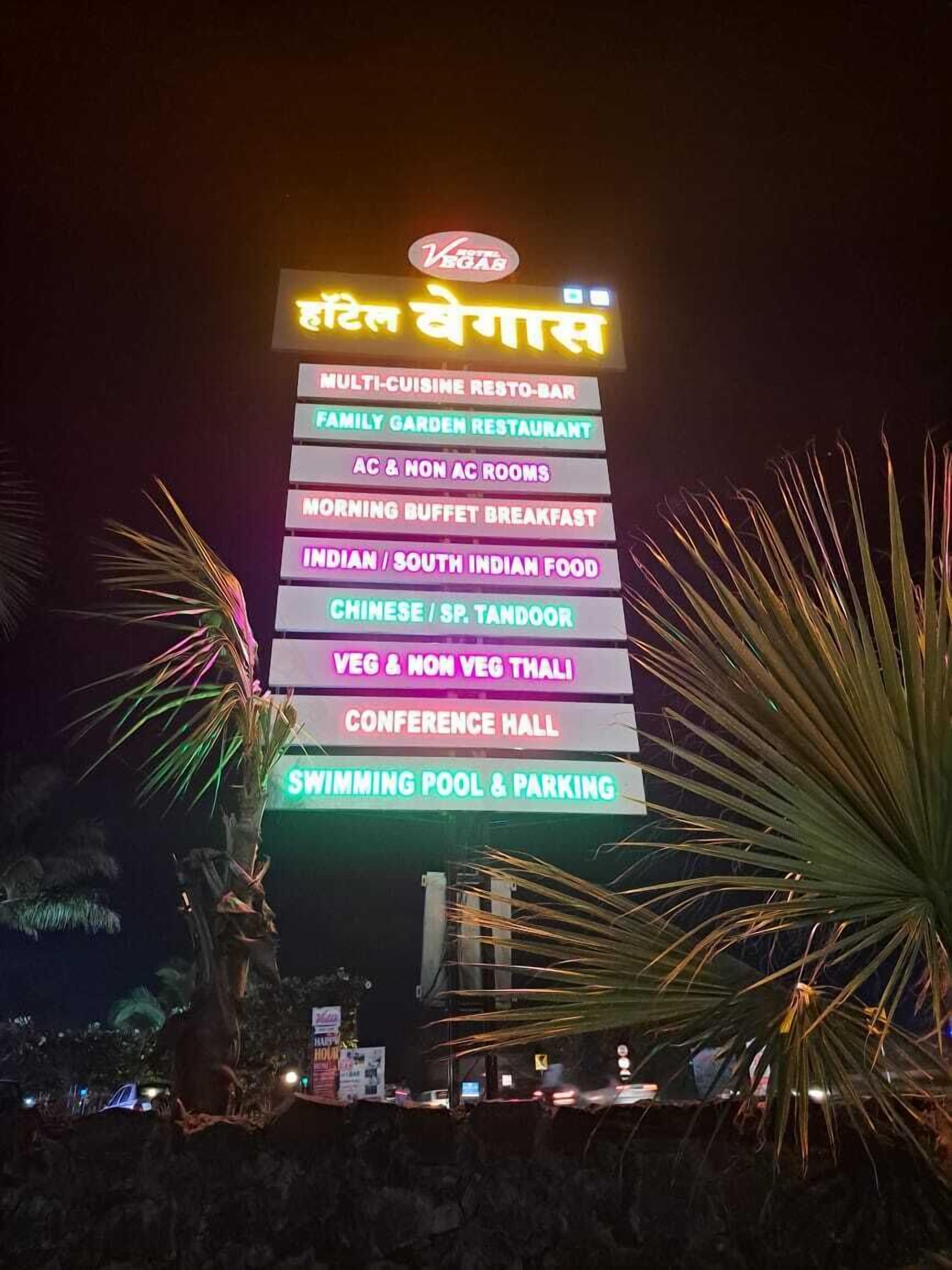 Un letrero de neón que enumera los servicios, incluyendo un restaurante de cocina internacional, restaurante jardín para familias, desayuno, comida india, comida china, opciones vegetarianas, salón de conferencias y piscina.