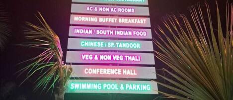 Un letrero de neón que enumera los servicios, incluyendo un restaurante de cocina internacional, restaurante jardín para familias, desayuno, comida india, comida china, opciones vegetarianas, salón de conferencias y piscina.