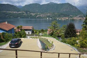 Vista costera con un automóvil estacionado en la entrada, un edificio de techo rojo y montañas al fondo.