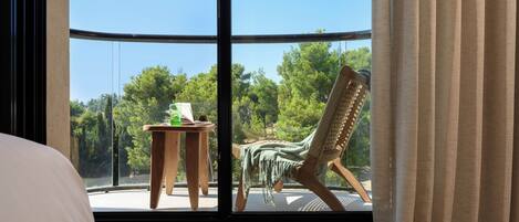 Una habitación con un ventanal grande, una cama, una silla de madera y una mesita con un objeto verde encima.