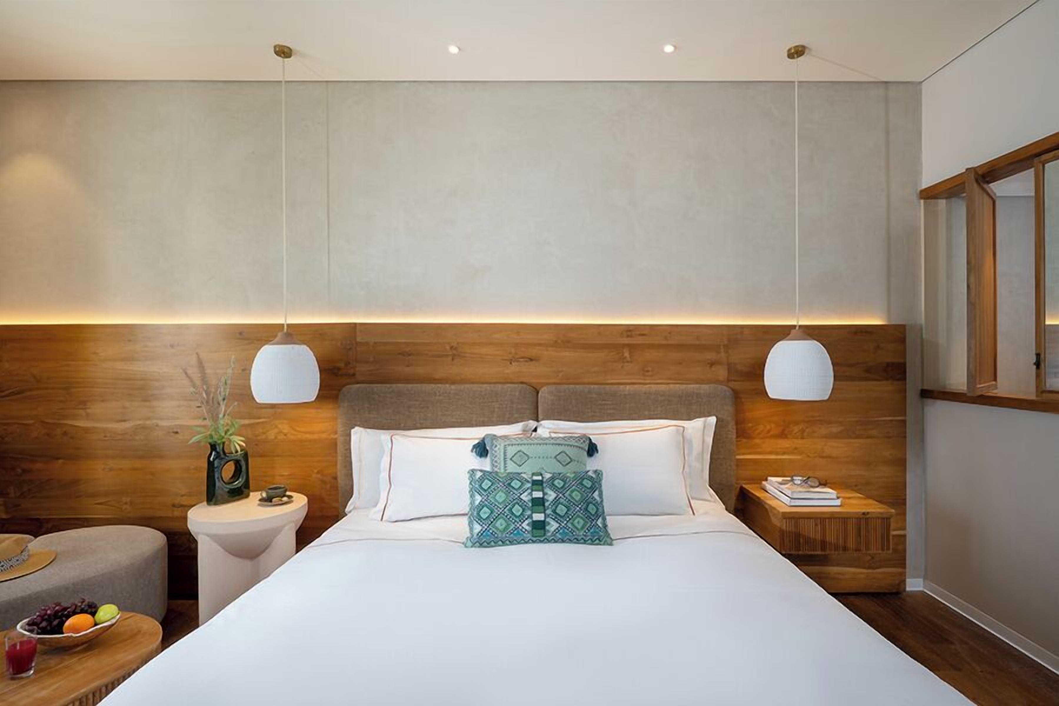 A modern hotel room with a wooden headboard, a large bed with white bedding, two bedside tables with lamps, a small round table with fruit, and a wooden bench.