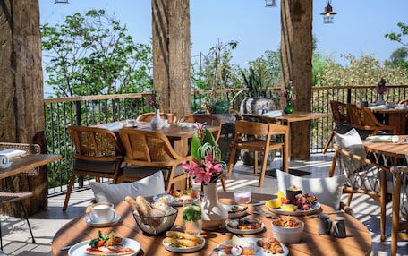 An outdoor dining area with wooden tables and chairs, surrounded by trees and plants.