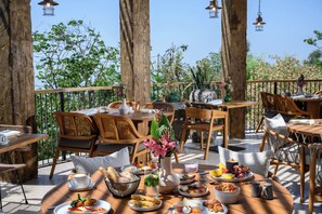 Un espace extérieur aménagé pour les repas, avec des tables et des chaises en bois, entouré d’arbres et de plantes.