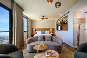 Una habitación de hotel moderna con un sofá curvo, una pequeña mesa redonda con un libro y un vaso de agua, una cama con cabecero de madera y vistas a la ciudad a través de ventanas grandes.