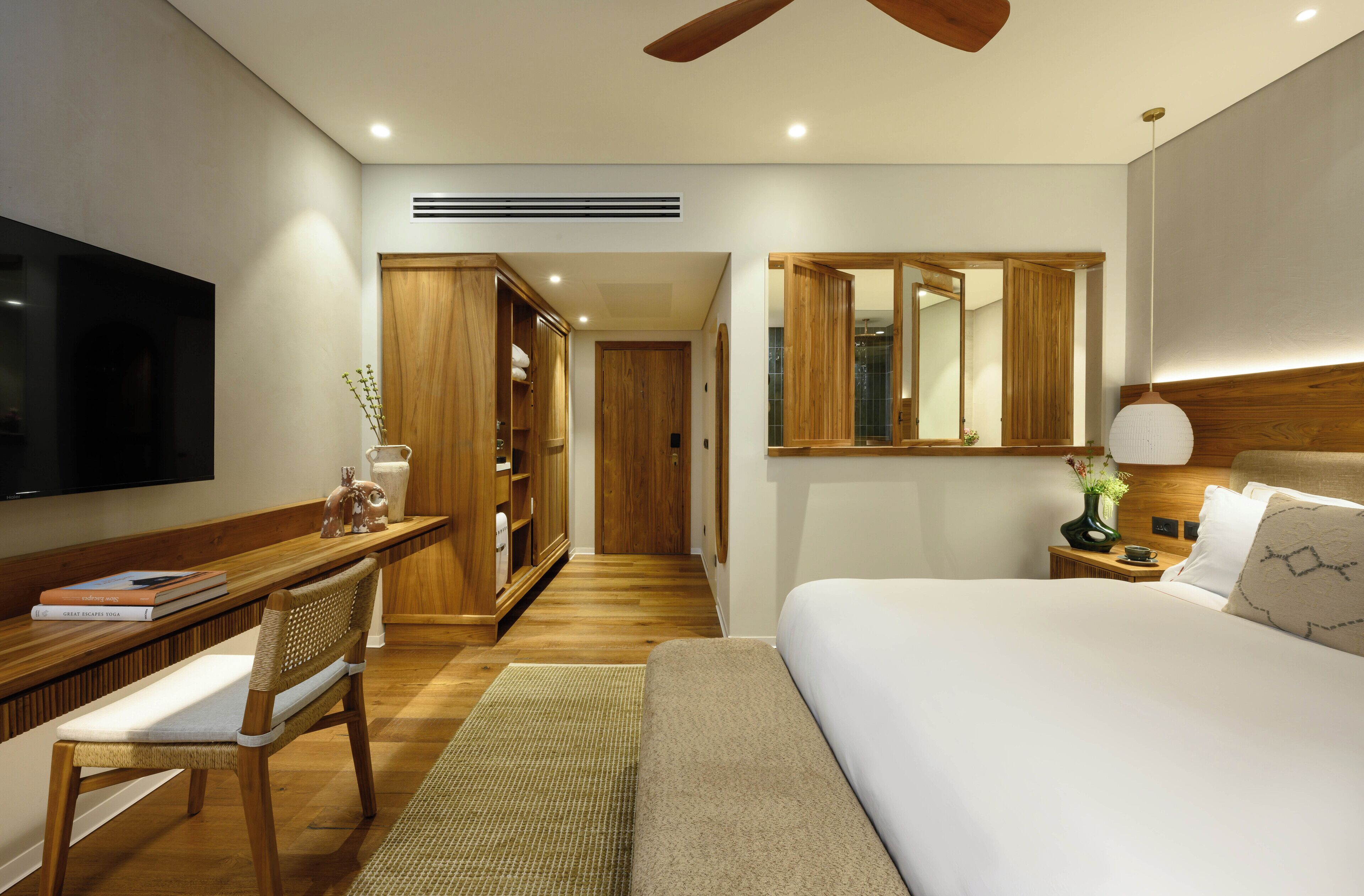 A modern hotel room with a large bed, a desk with a chair, a TV, a wardrobe, and a mirror.