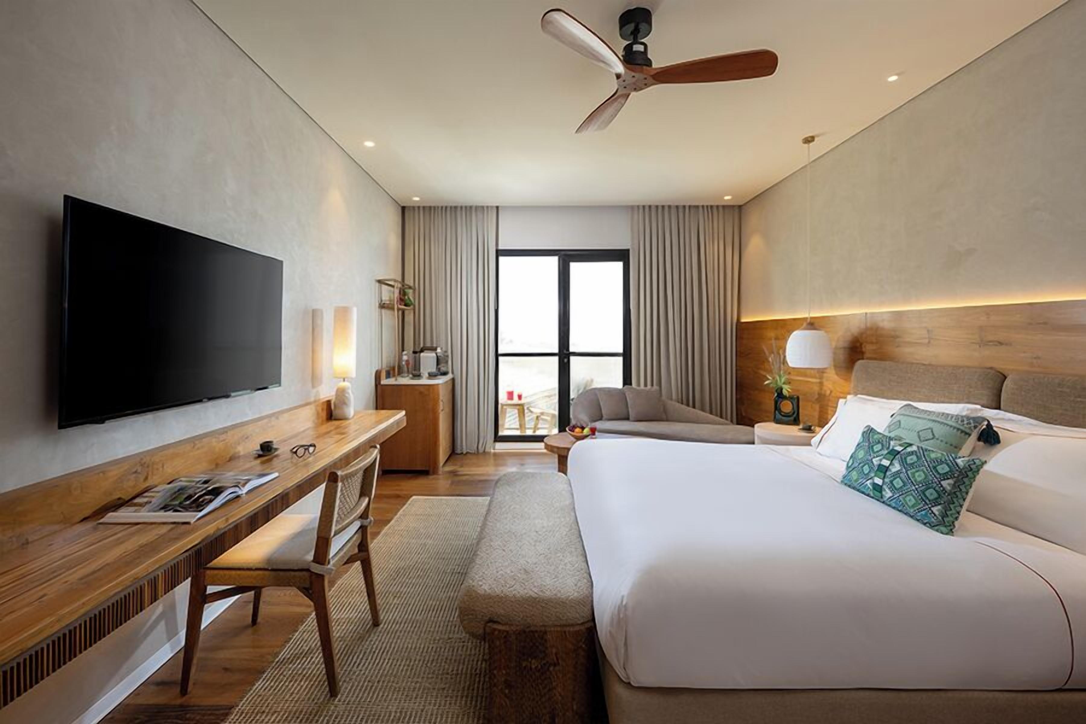 A modern hotel room with a flat-screen TV, a wooden desk, a comfortable sofa, a large bed, and a ceiling fan.