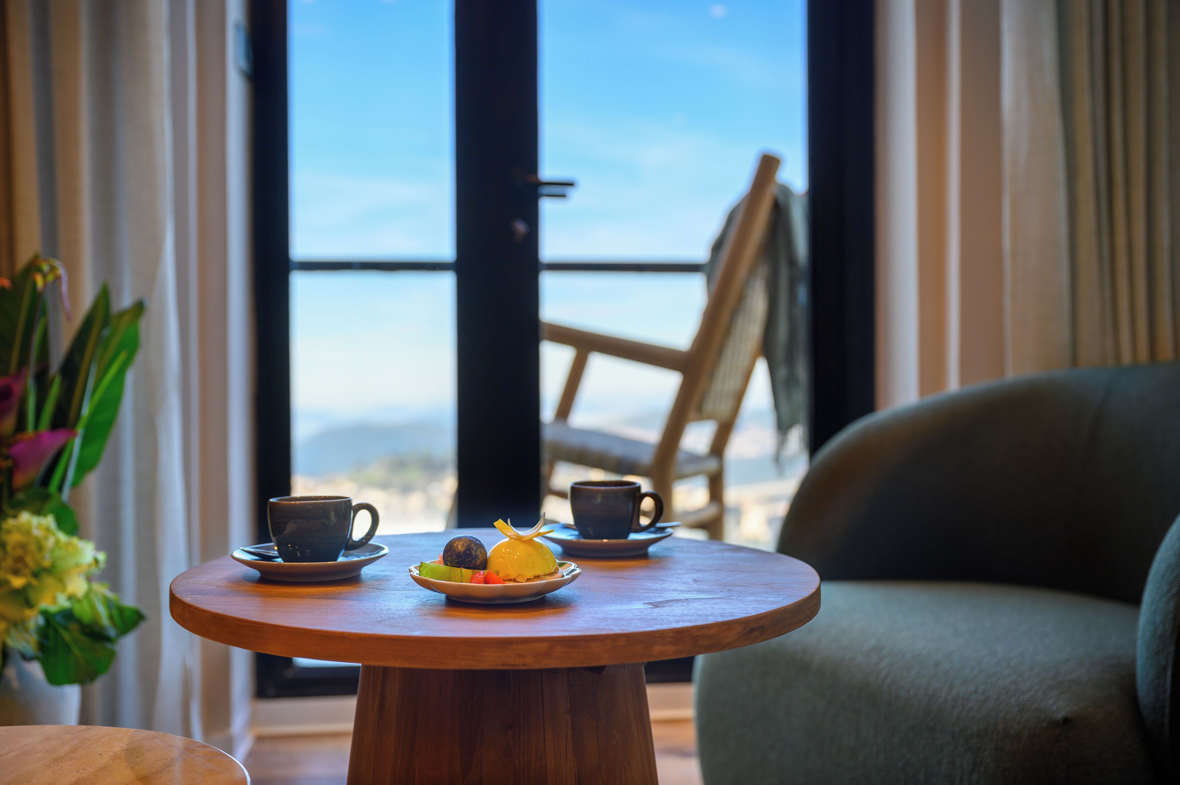 Una mesa de madera redonda con un plato de fruta y dos tazas, una silla, y vistas al exterior.