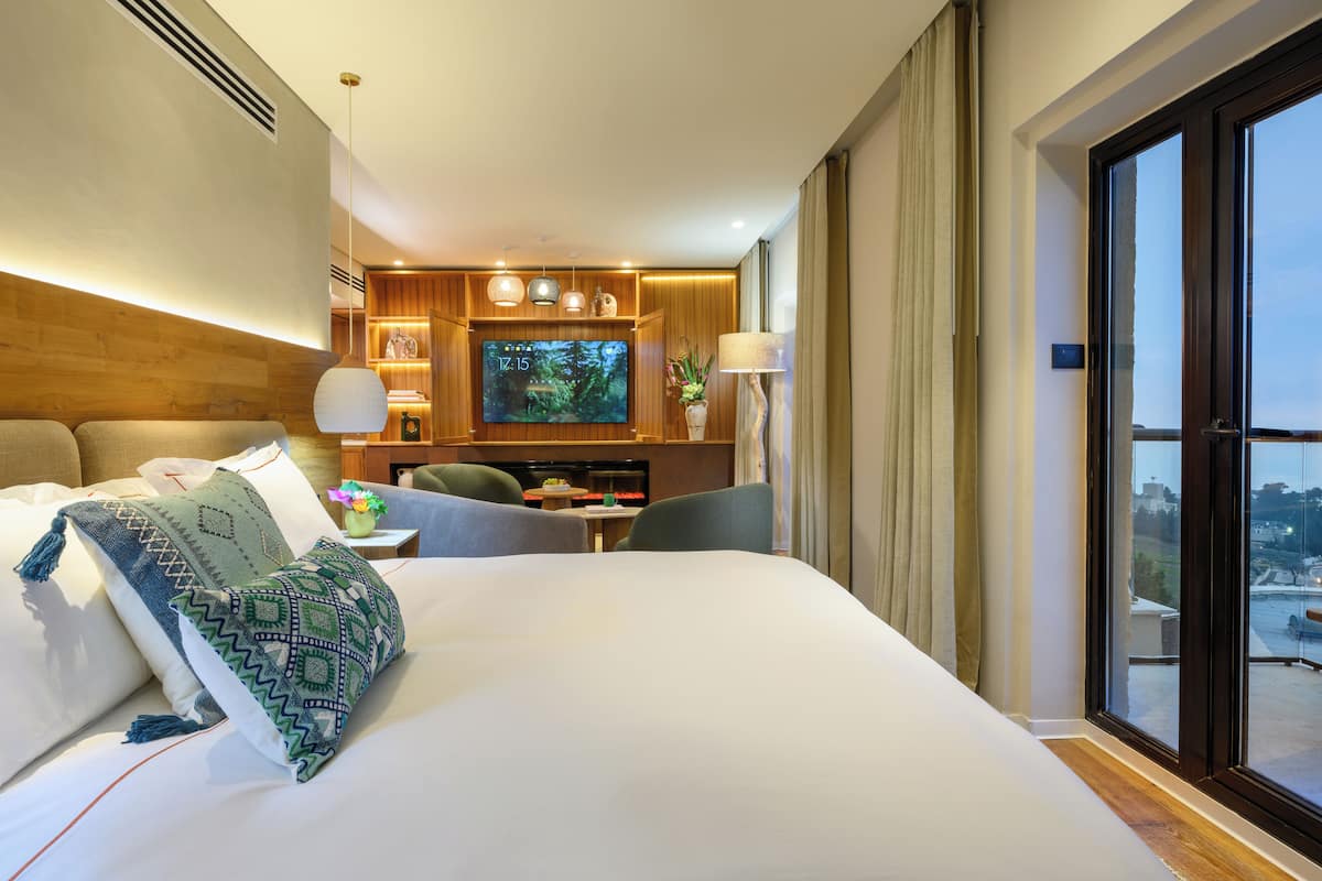 A hotel room with a large bed, a TV, a desk with a chair, and a view of the city through the window.