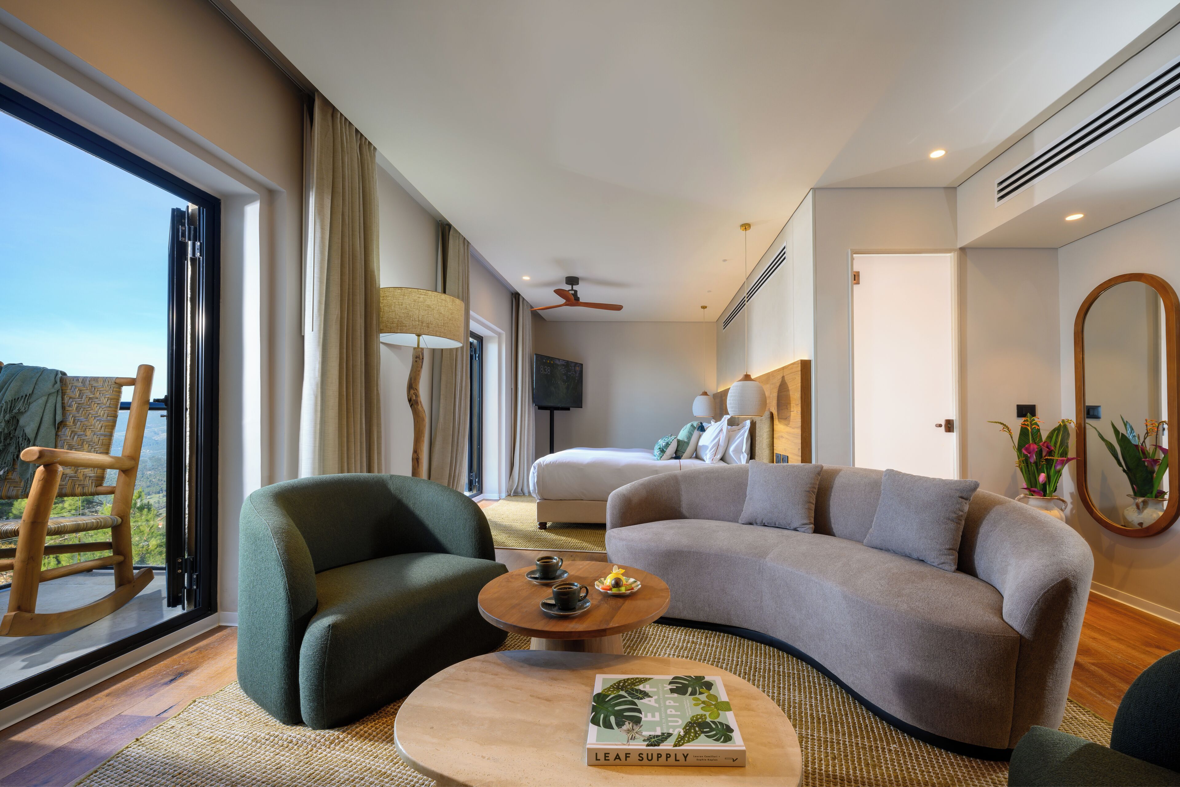 Habitación de hotel moderna con un sofá gris curvo, un sillón de madera mecedor, una mesita redonda con un libro y un ventanal con vistas.