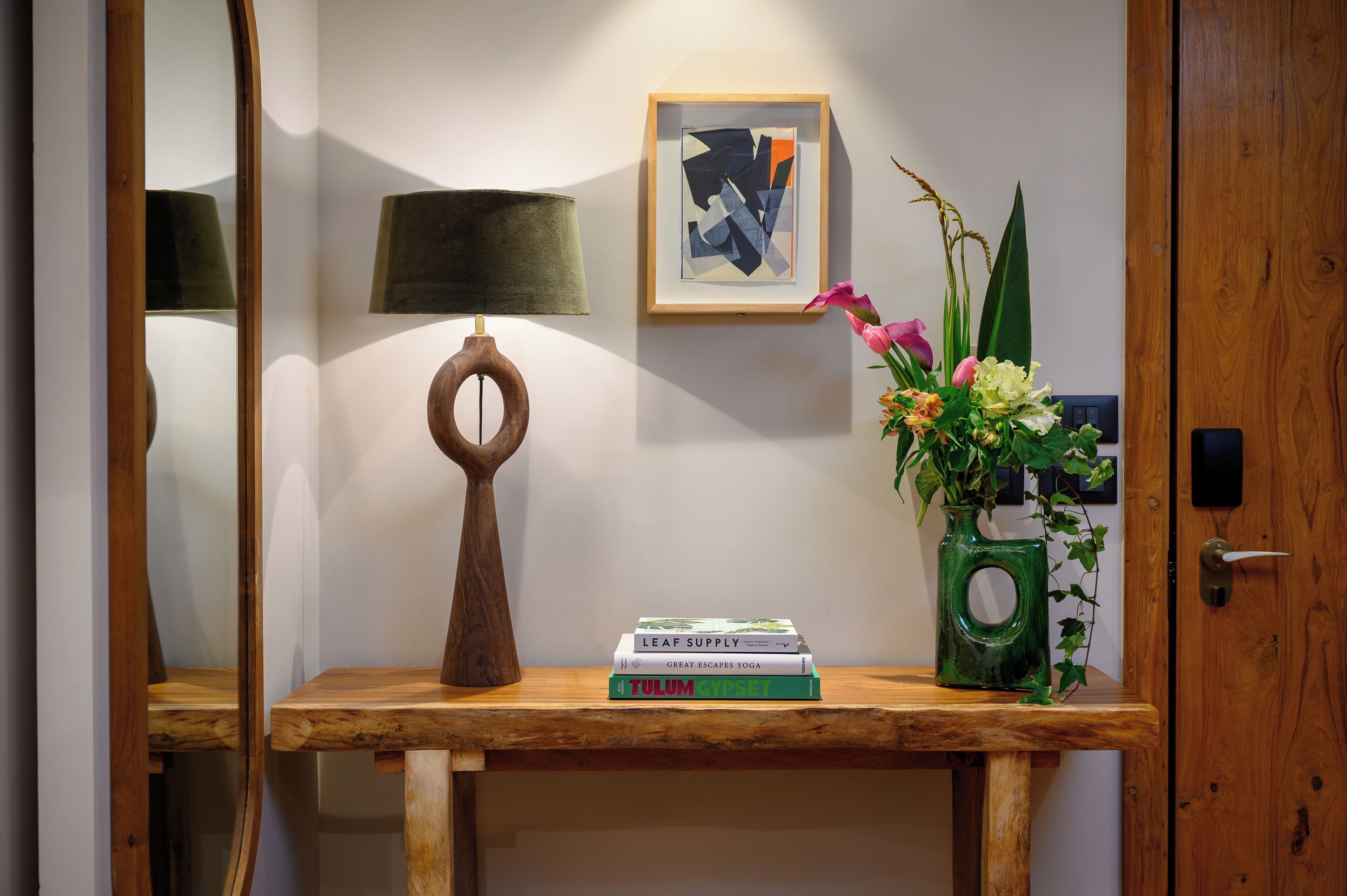 Una consola de madera con una lámpara, obras de arte enmarcadas, un florero con flores y una pila de libros.
