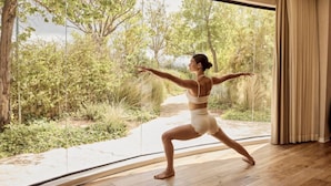 A person in a yoga pose in a room with a large window, wooden flooring, and a curtain. - Canaan Hotel - Limited Edition By Fattal (Safed)