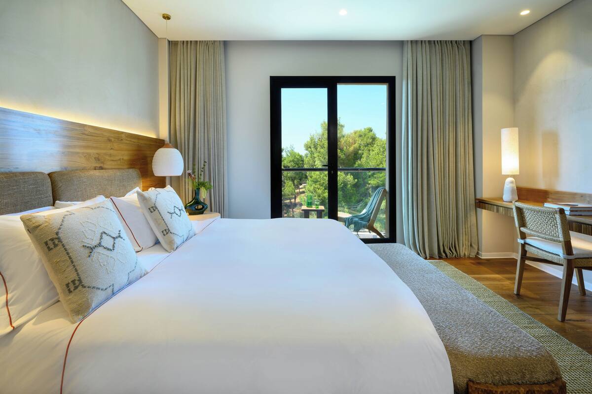 A hotel room with a large bed, a desk, and a view of greenery through the window.
