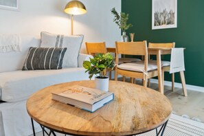 A cozy living room with a round wooden table, a sofa with cushions, a floor lamp, and a potted plant.