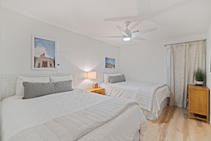 A hotel room with two beds, a ceiling fan, a nightstand with a lamp, a window with curtains, and a plant on a side table.