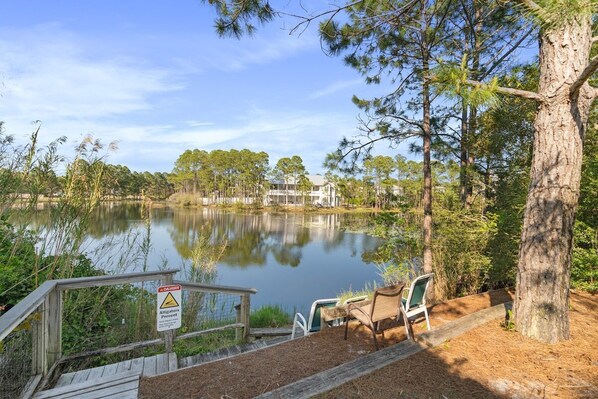 A lakeside area with a wooden deck and chairs, a warning sign for alligators, and a building in the background.