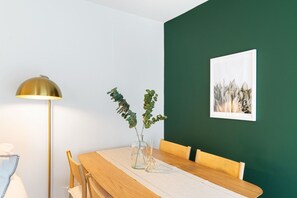 Ein Essbereich mit einem Holztisch, Stühlen, einer klaren Vase mit Grünpflanzen, einer Stehlampe und einem gerahmten Kunstwerk an der Wand.