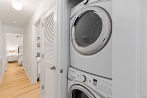 A laundry room with stacked washing machines and dryers, a wooden floor, and a hallway leading to a bedroom.