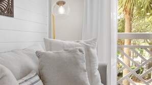Pared de madera blanca con un panel decorativo, una lámpara colgante de vidrio transparente y vistas a palmeras a través de una ventana de celosía blanca.