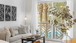 A modern living room with a sofa, coffee table, and a view of palm trees through large windows. - 🌴 Stylish Studio Retreat in Sandestin Resort (Miramar Beach)