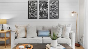 Una sala de estar con un sofá, una mesa de centro con libros y una planta, una lámpara y una pared con tres paneles decorativos.