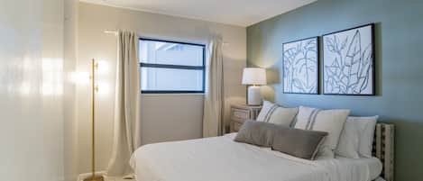 Un dormitorio con una cama, una mesita de noche, una lámpara, una ventana con cortinas y dos cuadros decorativos.