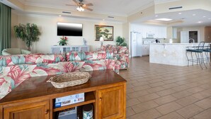 A spacious living room with a sofa, a wooden coffee table, and a TV. There is a kitchen area with white cabinets and a dining space with bar stools. - Phoenix West 1209 4 Bedroom Condo (Orange Beach)