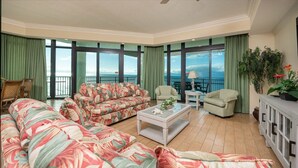 A spacious living room with a large sofa, two armchairs, a coffee table, and a view of the ocean through large windows. - Phoenix West 1209 4 Bedroom Condo (Orange Beach)