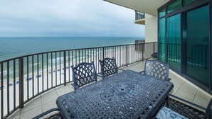 A balcony with a view of the beach and ocean, a table with chairs, and a glass door. - Phoenix West 1204 3 Bedroom Condo (Orange Beach)