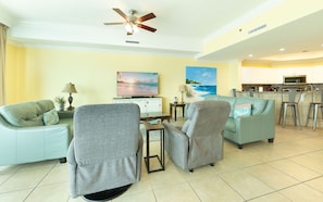 Sala de estar com dois sofás, televisão, ventilador de teto e área de cozinha com micro-ondas.