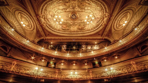 Interior de un teatro ornamentado con un techo imponente, varios balcones y elaboradas arañas de cristal.