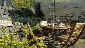 Juego de mesa y sillas de madera al aire libre, frente a una pared de piedra, con una escalera y una lámpara colgante cerca.