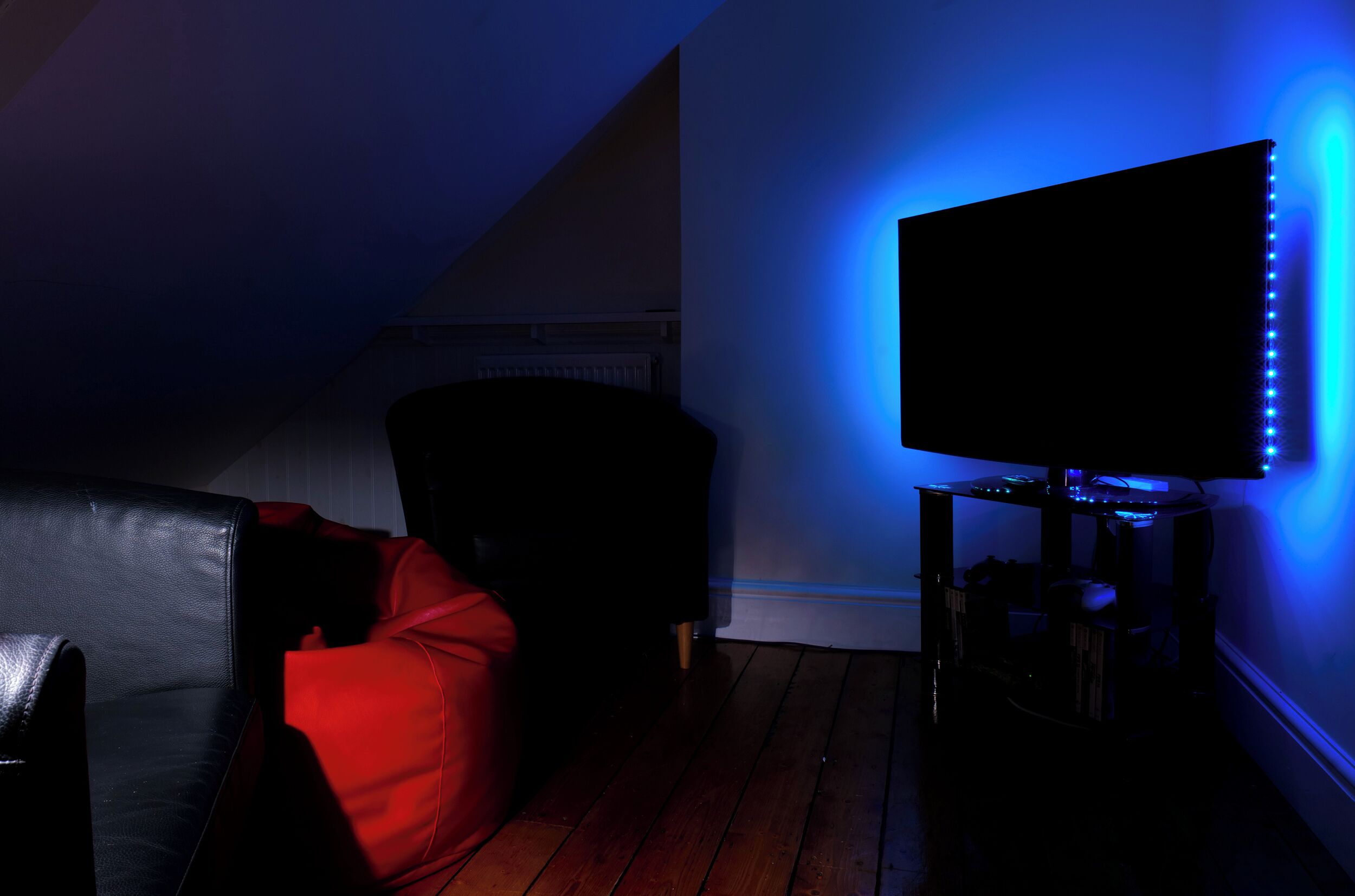 A room with a black leather sofa, a red chair, and a TV mounted on the wall with blue LED lights.