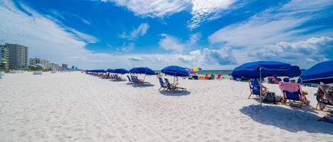 A beach with rows of blue umbrellas and lounge chairs, a clear sky, and buildings in the background.