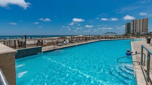 Une piscine à l’eau d’un bleu cristallin, entourée de chaises longues et de parasols, avec une plage et l’océan en arrière-plan.