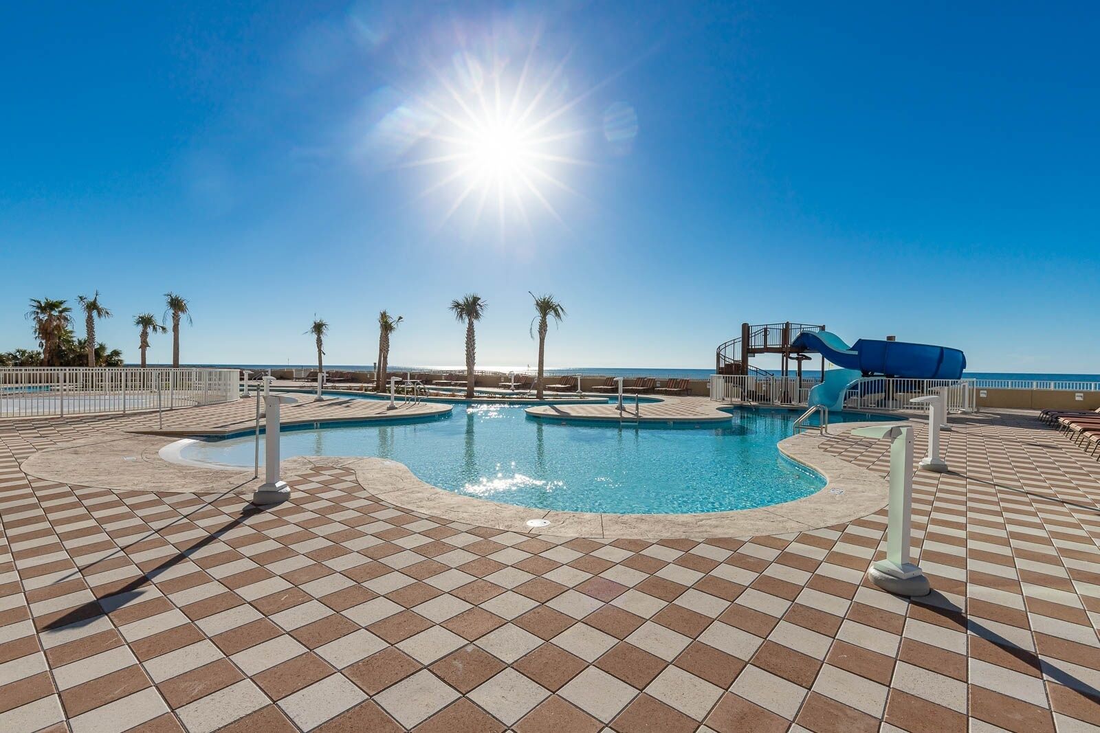 A swimming pool with a water slide, palm trees, and a clear blue sky.
