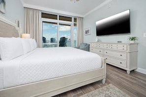 A bedroom with a large bed, a dresser, a flat-screen TV, and a view of the outdoors.