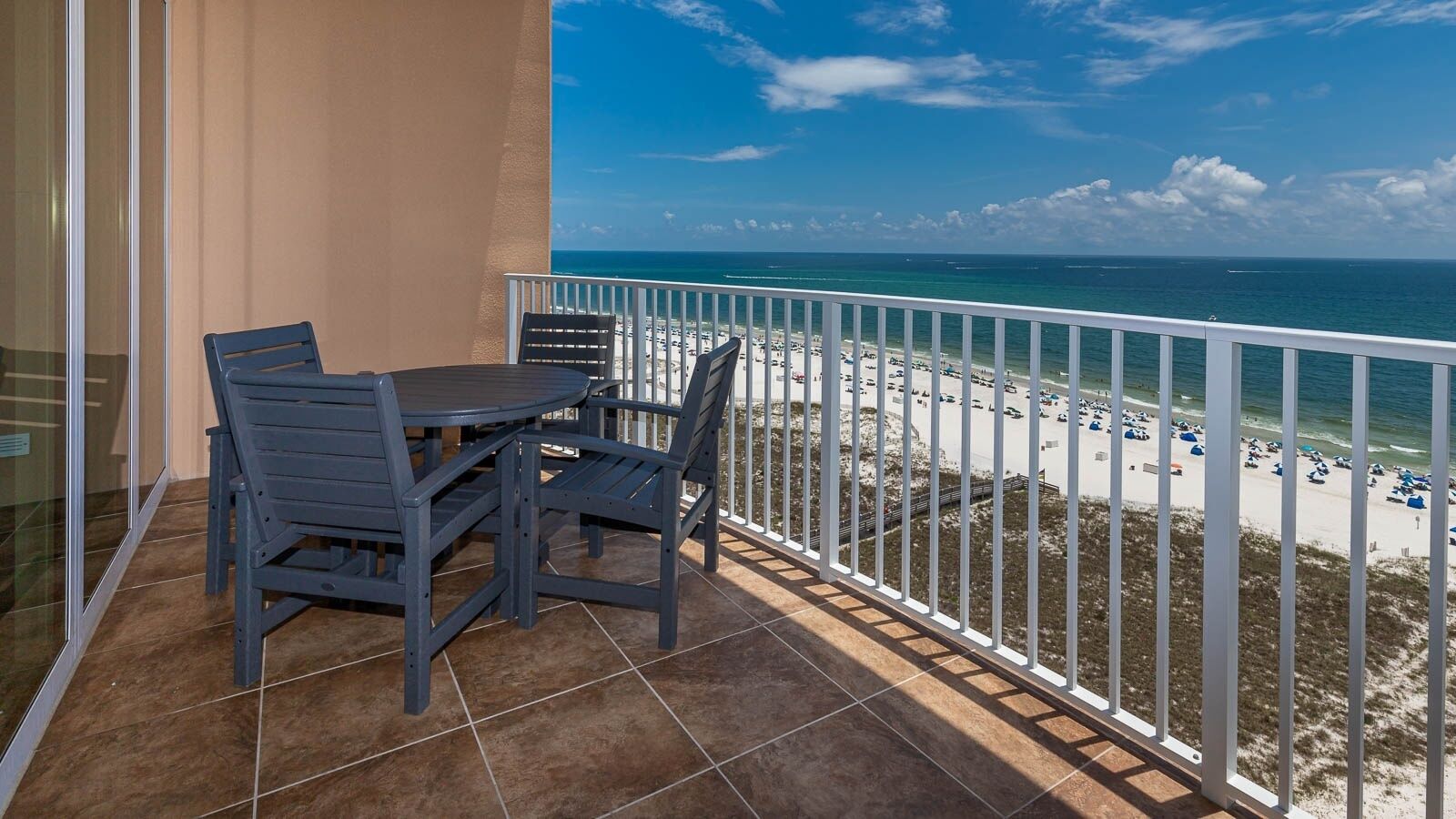 A balcony with a table and chairs, overlooking a beach with umbrellas and a clear blue sky.