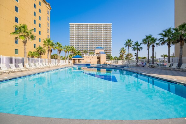 A swimming pool with lounge chairs and palm trees, surrounded by a fence, with a tall building in the background. - Crystal Tower 2004 2 Bedroom Condo (Gulf Shores)