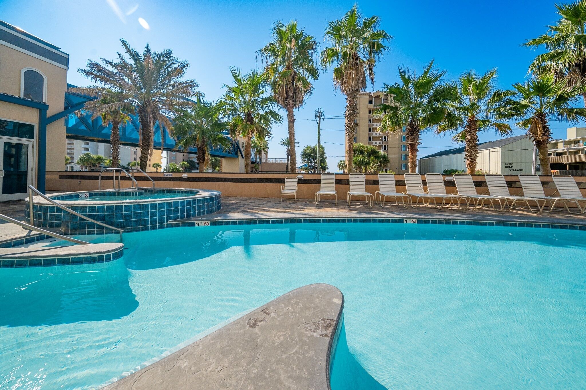 A swimming pool with lounge chairs, palm trees, and a building in the background.