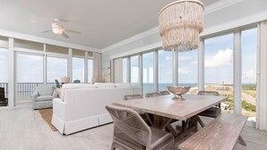 A spacious room with a large window, a ceiling fan, a dining table with chairs, and a white sofa. - Phoenix Gulf Towers 1101 4 Bedroom Condo (Orange Beach)