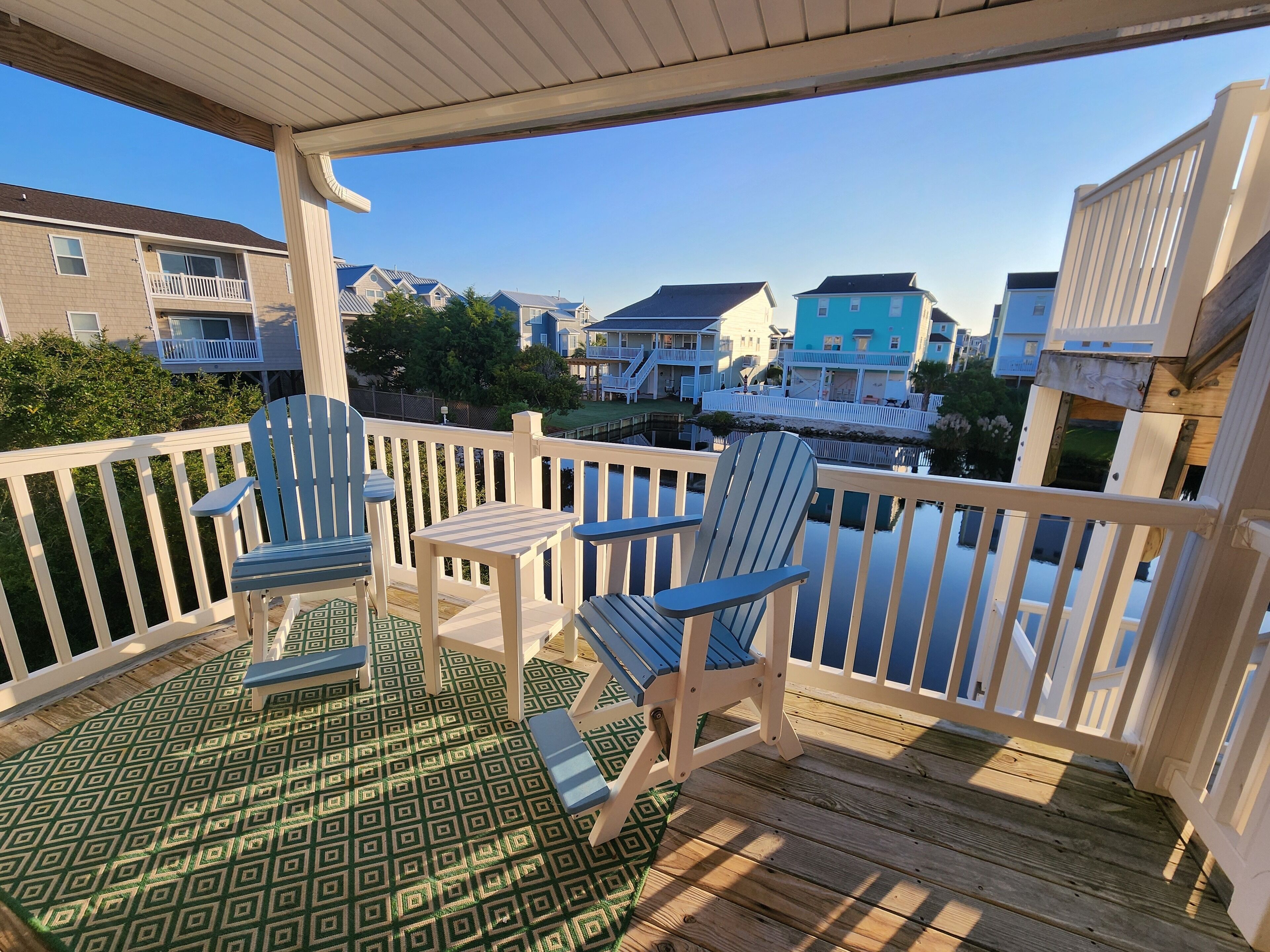 A wooden deck with two Adirondack chairs and a small table, overlooking a residential area with houses and a body of water.