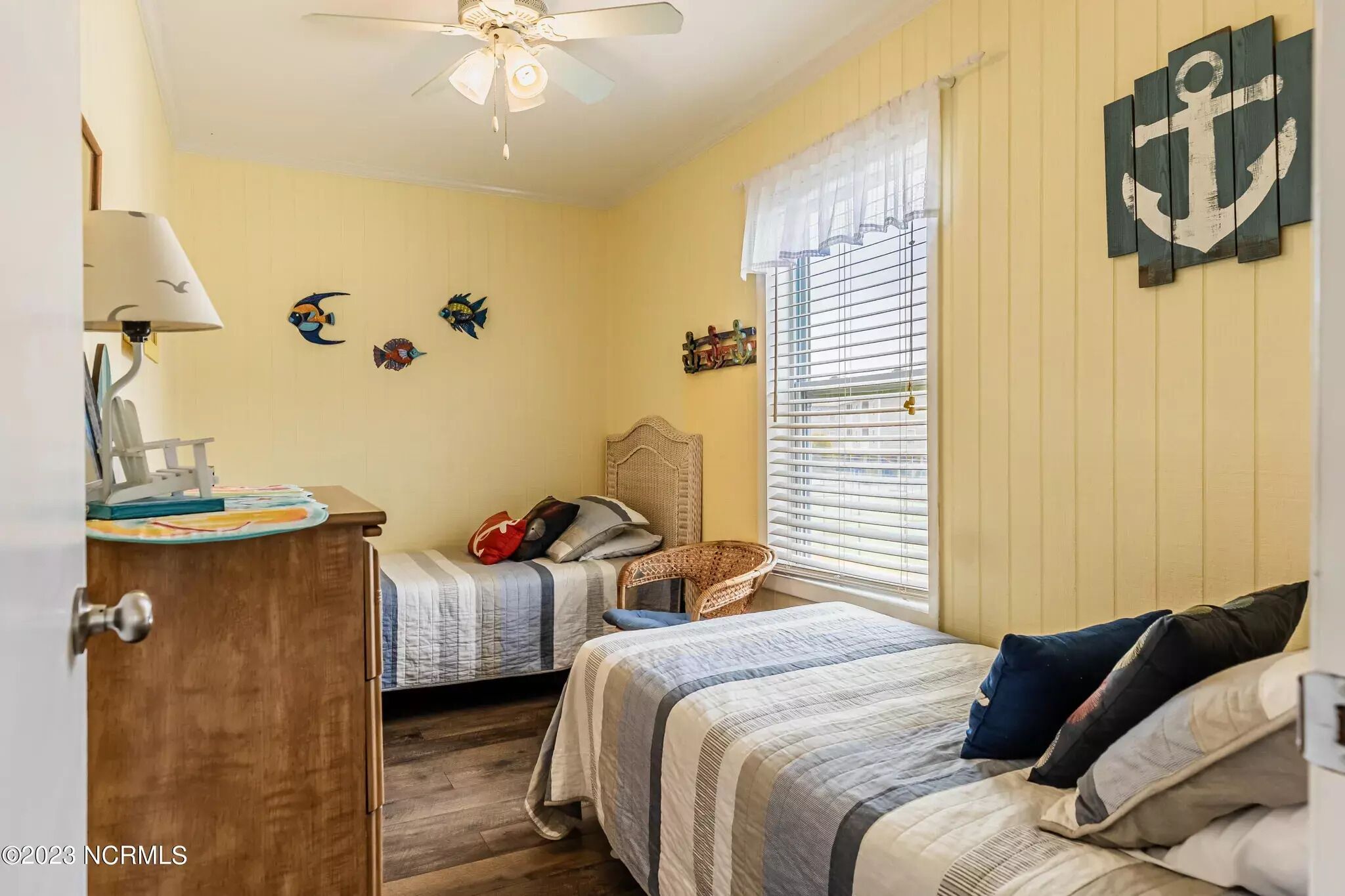 A bedroom with two beds, a ceiling fan, a window with blinds, and wall decorations including fish and an anchor.