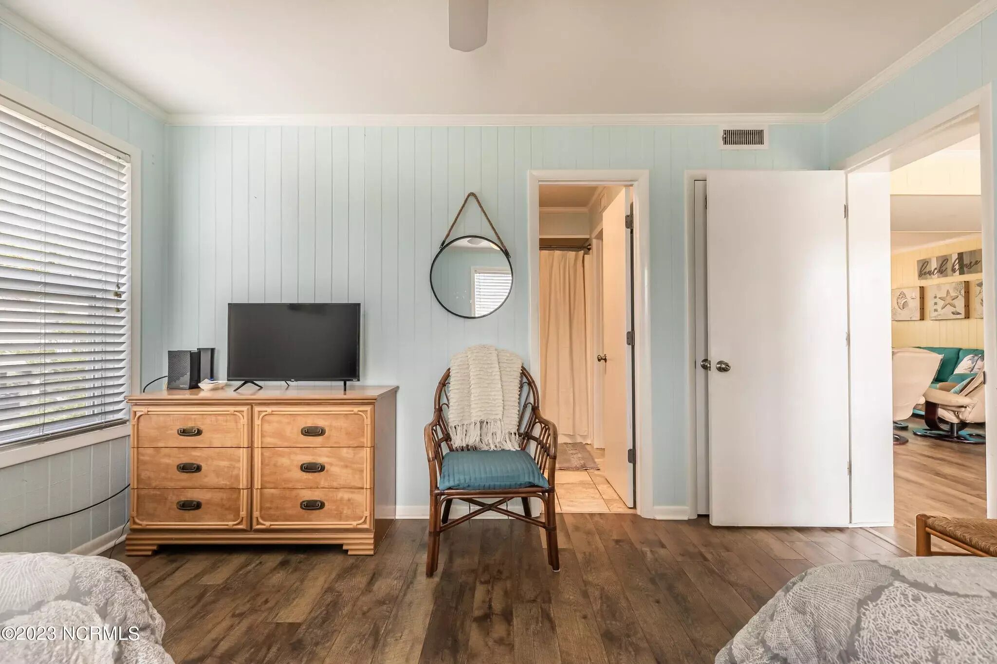 Une chambre à coucher avec une commode en bois, un téléviseur, une chaise et une porte donnant sur une autre pièce.