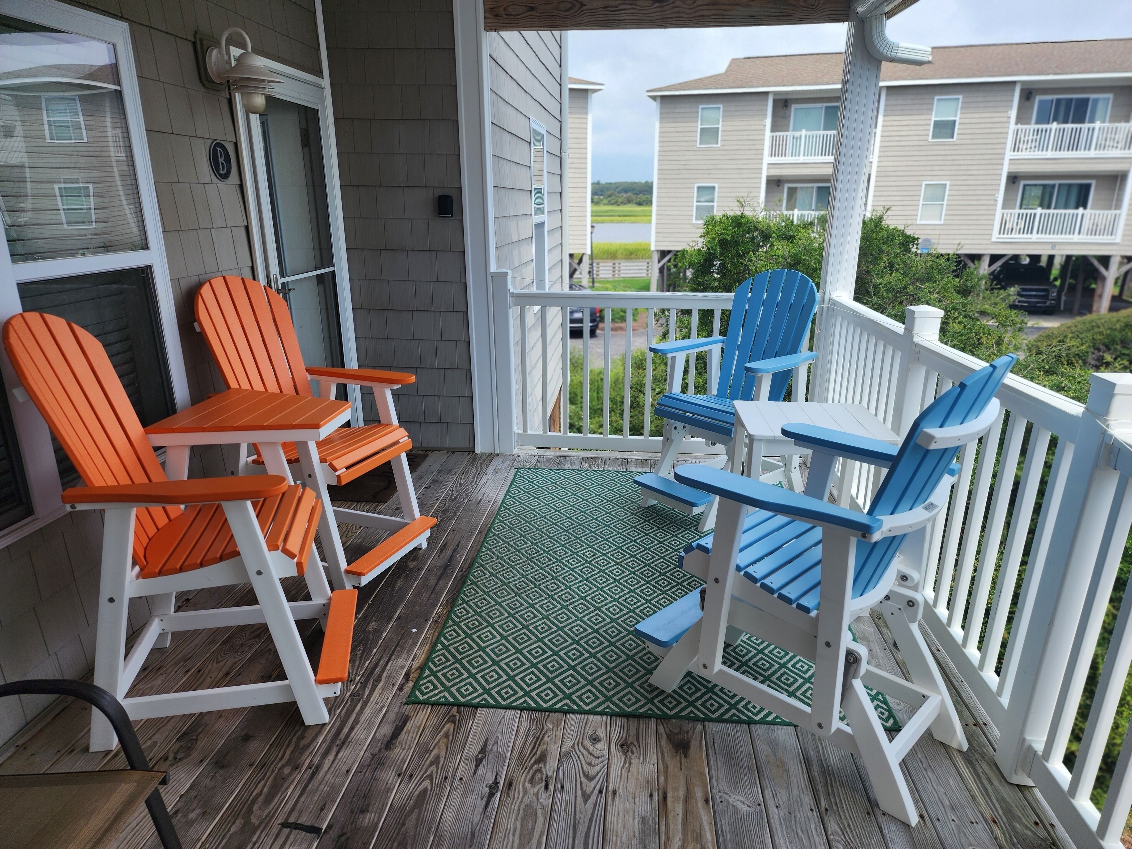 A covered porch with two sets of Adirondack chairs, one set orange and one set blue, and a green patterned rug.