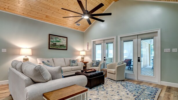 A spacious living room with a ceiling fan, a large window, and a view of the outside deck.