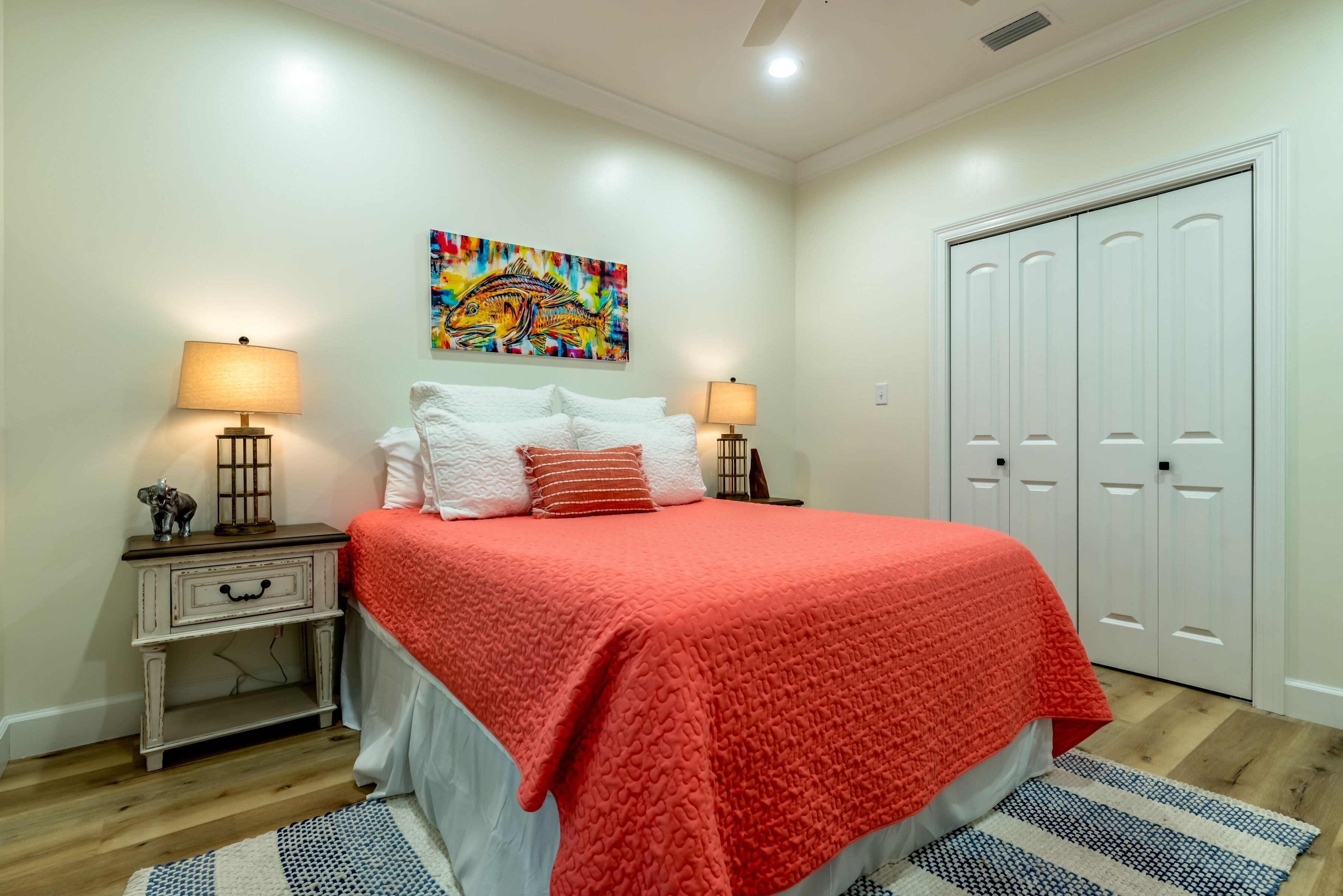A bedroom with a bed, two bedside tables, a nightstand, a colorful painting, and a white wardrobe.