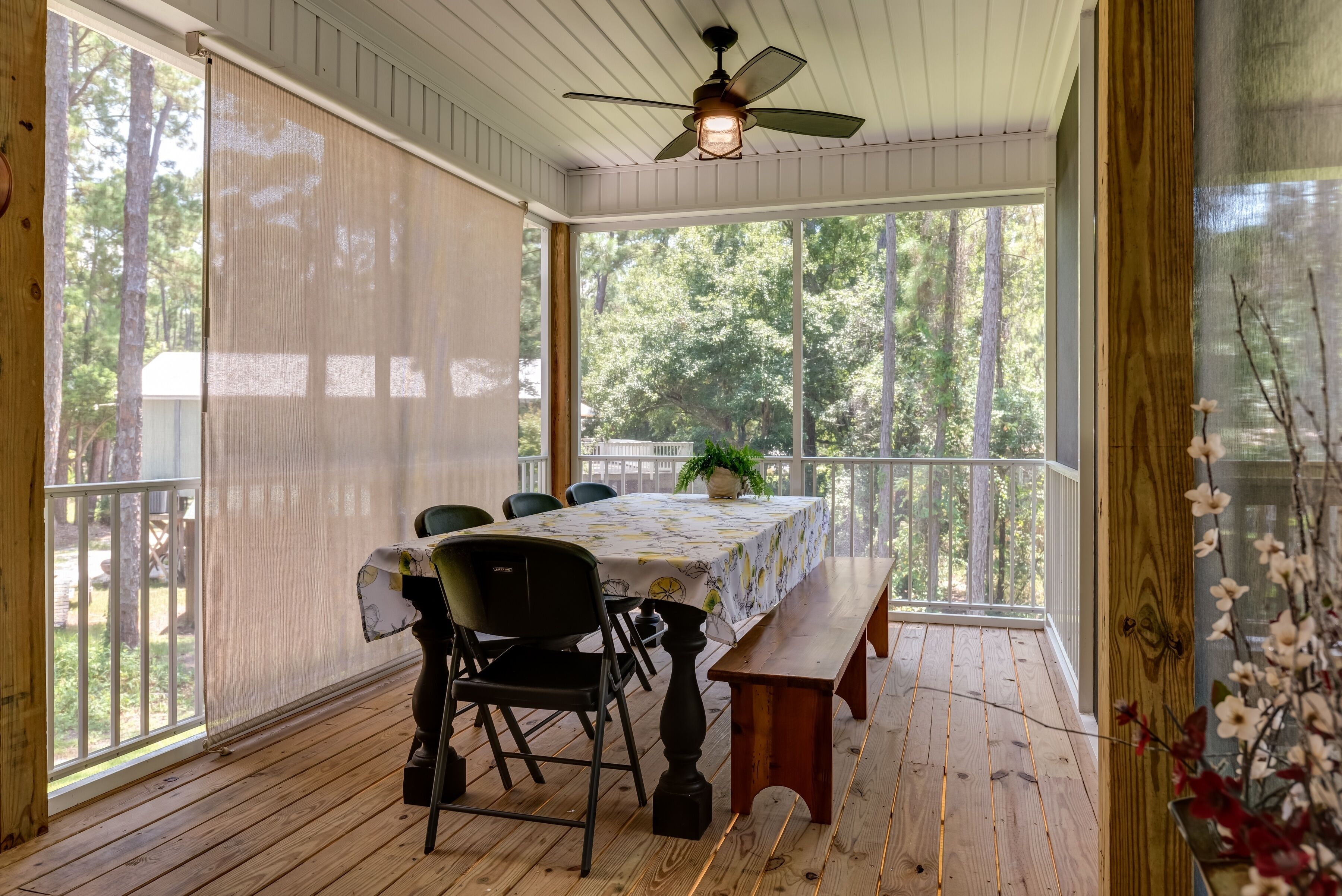 A covered porch with a wooden table and chairs, a ceiling fan, and a view of trees through large windows.